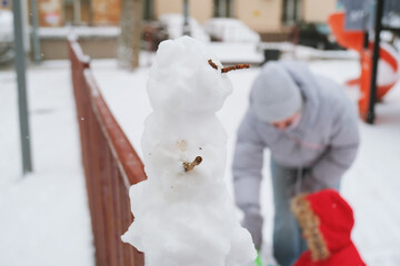 Snowy day at a park features a small snowman on a fence with a child in a red coat playing nearby....