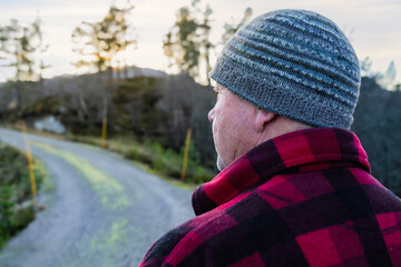 Man wearing a knitted hat and red plaid shirt stands on a winding gravel road, surrounded by trees, reflecting on nature and the beauty of the outdoors in a serene environment