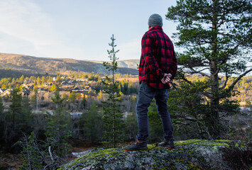 Male hiker in a red plaid shirt stands on a rock, gazing at a scenic landscape with trees and mountains, embodying adventure and exploration in nature