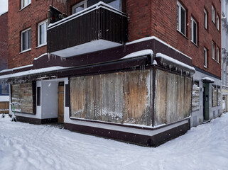 A snowy urban street scene featuring a corner brick building with boarded-up windows and a closed ground-floor storefront.