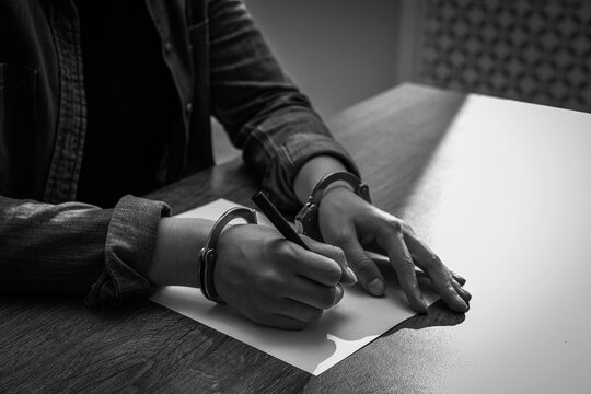 Handcuffed woman writing confession at table, closeup. Captured criminal. Black and white toning