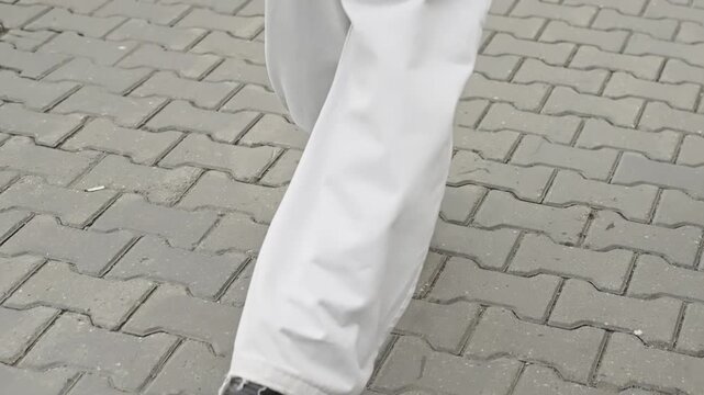 Close-up view of a pedestrian's feet in black shoes and loose light-colored trousers walking across a patterned stone sidewalk, capturing motion and urban details on pavers