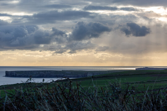 Dramatic coastal landscape at sunset with sunbeams breaking through storm clouds. A scenic view of green fields and rugged cliffs overlooking the shimmering ocean in the British countryside in Wales - Powered by Adobe