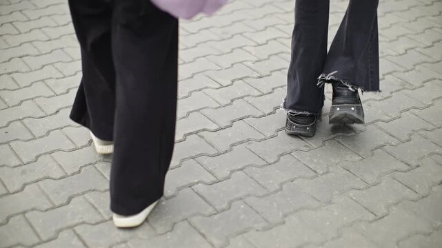 Close-up rear view of two people walking side by side on an urban paved sidewalk, showing moving legs, flared trousers, and contrasting footwear stepping across interlocking tiles.