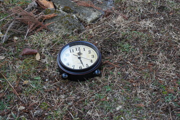 A black clock with a white face sits on the ground in a grassy area. The clock is set at 3:00