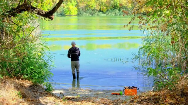 Man in boots with fishing rod on bank of autumn river. A fisherman catches pike, perch or carp in calm water. View from back. Nature adventures, relaxing holidays, fishing hobby concept