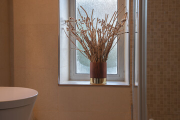 Minimalist dried flower arrangement in a brown and gold vase on a bathroom window ledge, adding...