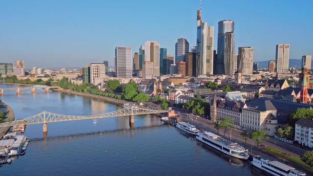 Aerial drone zoom-in shot from 100m over the Main River towards the Eiserner Steg bridge and the modern skyscraper skyline. Scenic urban landscape of the financial district in Frankfurt, Germany.