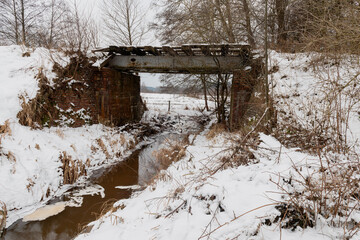 Strumień płynący pod starym mostem kolejowym. W poprzek strumienia jest tama z gałęzi zbudowana przez bobry. Jest zima, brzegi i tamę pokrywa warstwa  śniegu.