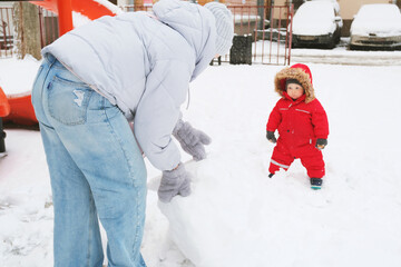 Child in red snowsuit watches an adult assembling a snowball in a snowy park. The child looks excited by the playful activity, creating a joyful winter moment.