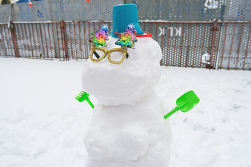 A whimsical snowman adorned with colorful decorations and a bucket hat, holding green tools in a snowy playground. Bright sunglasses add a playful touch to this winter scene.