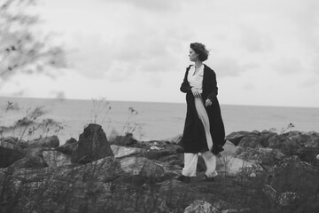 Naklejka premium Woman on a rocky coast in a long coat walking along the shore, black and white scene with wind swept hair and calm sea horizon