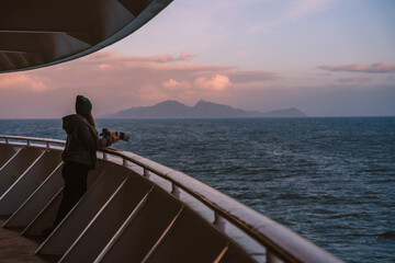 Fototapeta premium Female Photographer on Cruise Ship Looks At Mountain at Sunset. Adventure Travel. Beautiful Girl On Deck Handrail Balcony Looks Over Ocean