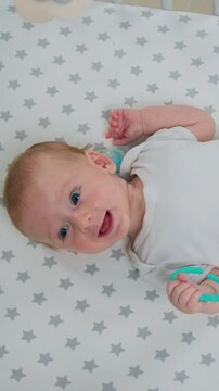 Portrait of smiling adorable baby boy lying on back and actively moves his arms and legs in baby cot. Close up happy 2 months old infant playing with teether of teeth in crib, top view. Slow motion