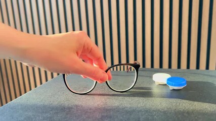 A woman’s hand places a contact lens case on a table, then sets down her eyeglasses, showing eye care routine and vision correction essentials in close detail.