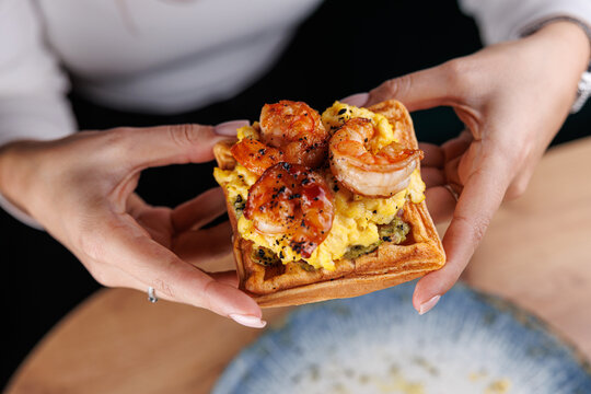 Unique dish with shrimp and scrambled eggs served on a waffle by a person at a dining table in a casual restaurant setting