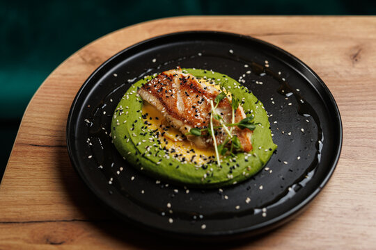 Fusion dish featuring grilled fish atop green puree with sesame seeds, served in a modern restaurant setting