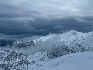 Obraz premium Dark winter storm clouds hang over a rugged mountain range covered with fresh snow. Mist rolls through the valleys, adding to the whimsical alpine atmosphere. Dramatic views in snowy Slovenian Alps.