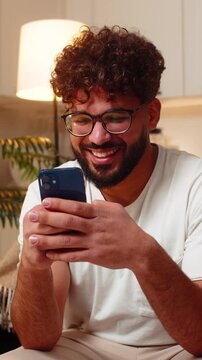 Arabian man at home checks smartphone gets a message from friend smiles types reply feels joy. Young guy on sofa writes friendly text reply continues chat in messenger feels connected happy alert.