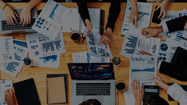 A diverse group of professionals engaged in a business meeting around a table filled with documents, charts, and laptops, promoting teamwork and data-driven decision-making. Raster - Powered by Adobe