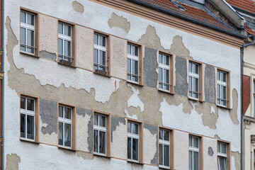 Building Facade With Peeling White Stucco Revealing Tan And Grey Underlayers Surrounding Two Rows Of Identical Rectangular Glass Paned Windows