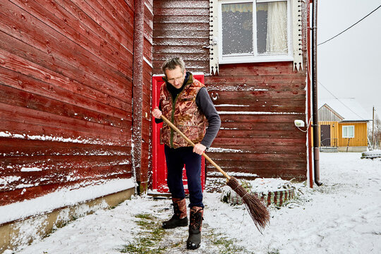 Homeowner sweeps snow from entrance path with birch broom, standing in front of rural house clad with painted red boards, wearing warm vest and winter boots.