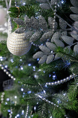A white textured Christmas ornament and glittery silver leaves on a pine branch with soft blue bokeh lights.
