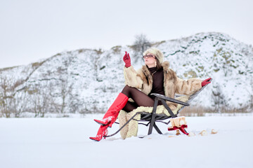 A chic woman in a luxurious fur coat and striking red boots finds solace in a chair amidst the winter landscape, savoring the serene beauty of the snowy scenery and exuding grace