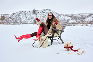 A stylish woman in a fur coat and red boots relaxes in a chair surrounded by a winter landscape and...