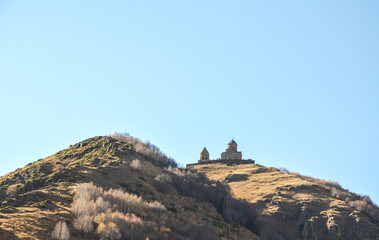Historic stone Gergeti Trinity Church stand atop a grassy, sunlit hill against a clear blue sky, with sparse trees covering the slopes below. One of the most famous landmarks in Georgia