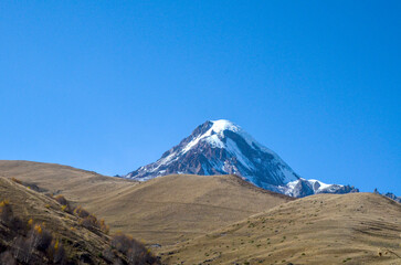 Majestic, snow capped Kazbek mountain peak rises sharply into a clear blue sky, seen from behind dry, golden brown foothills. Caucasus Mountains, Georgia