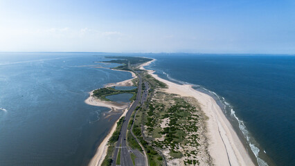 Aerial view of a long coastal barrier beach with a roadway, sand dunes, and blue ocean on both sides under a clear summer sky.