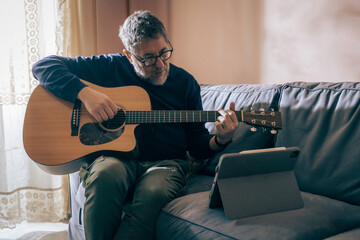 Obraz premium Man practicing guitar on sofa with tablet at home