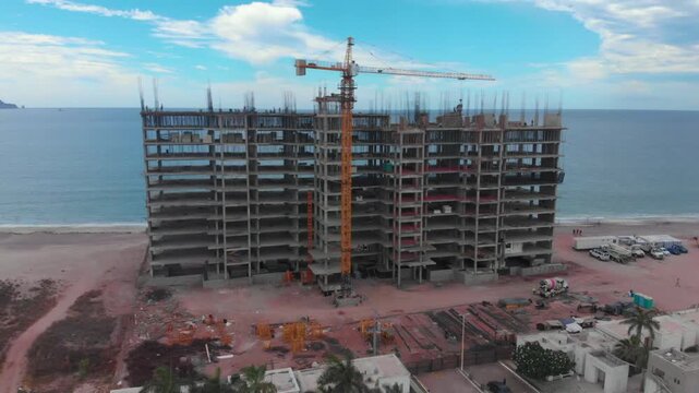 Toma de drone media luna de un edificio en construccion a la orilla del mar bajo un cielo azul con nubes blancas