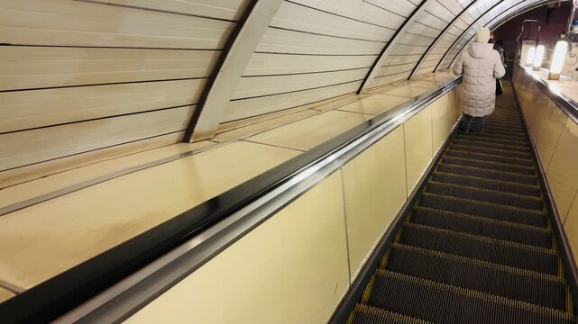 person on escalator in metro tunnel, arched perforated ceiling, geometric tread pattern, yellow paneling, solitary figure in light coat walking toward distant platform, leading