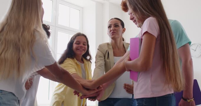 Happy school children group standing with their female friendly teacher or psychologist putting hands in a stack after successful therapy session in classroom. 4k video. Slow motion video.
