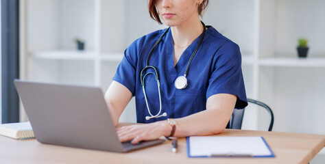A doctor or nurse in a medical uniform with a stethoscope sitting at a table with a laptop taking...