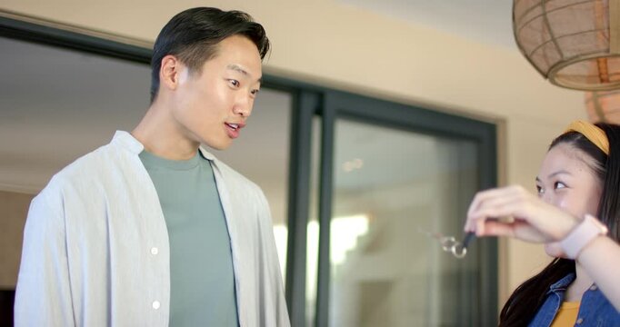 Asian couple exchanging smiles by sliding glass door, woman raising keys, man showing astonishment
