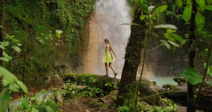 Woman walks on a mossy log near a powerful waterfall and a bright turquoise pool in the jungle