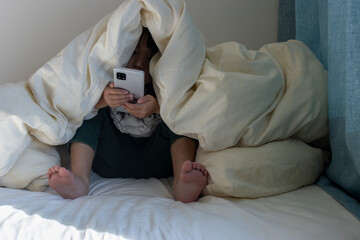 うす暗い部屋で布団をかぶってスマートフォンを見る女の子のイメージ（5歳、日本人、顔無し）