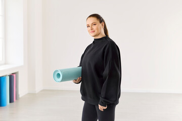 Yoga mat held in studio by woman. She stands in a minimalist room, holding a rolled mat, focused on...
