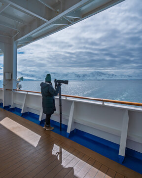 Female Photographer Antarctica Cruise Ship Holding Camera, Tripod Looking Over Handrail Aft Deck of Boat. Sunning Day Mountains Snow Glaciers Travel
