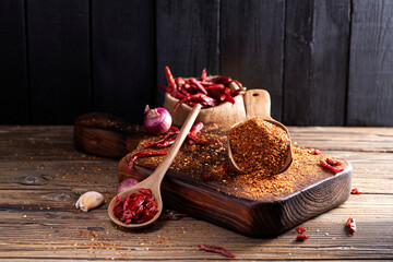 Chili powder in a wooden bowl and with a wooden spoon, along with garlic, shallots, and a mix of Asian spices, are arranged on a wooden table, conveying the concept of spicy food.
