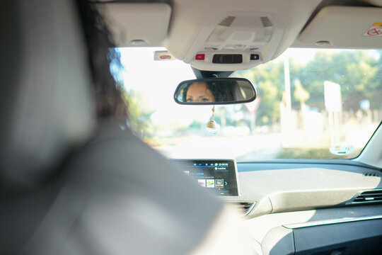A woman is driving a car and looking at her rearview mirror. The mirror has a small charm hanging from it - Powered by Adobe