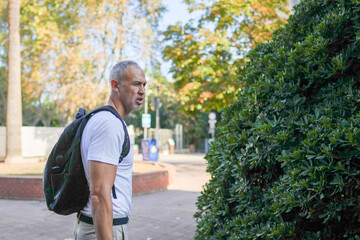Obraz premium A man wearing a backpack is standing in front of a bush. He looks like he is lost or confused