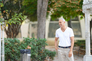 A man in a white shirt and khaki pants is smiling as he walks through a park. The scene is peaceful...