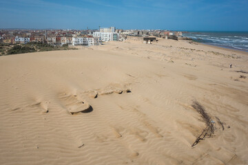 Panoramic view of the outskirts of Essaouira, Morocco, where the city meets vast coastal sand dunes. Residential buildings stand near the Atlantic shore under a clear blue sky