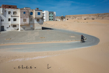 Edge of Essaouira, Morocco, where sandy dunes meet residential buildings. A lone cyclist rides along a narrow road bordered by sand and worn urban architecture under a clear sky