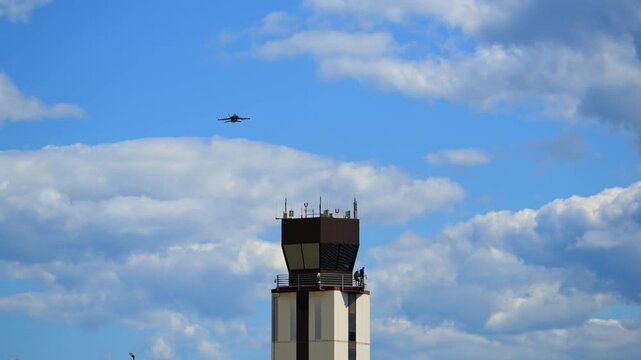 Military fighter jet fly past an airport control tower against a blue sky with drifting clouds, creating a dynamic aviation scene with airbase infrastructure. Bottom view