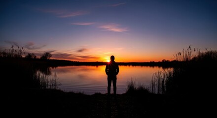 Solitary figure observes vibrant sunset reflected upon calm water surface